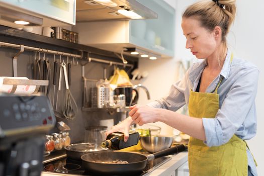 Stay at home housewife woman cooking in kitchen, stir frying dish in a saucepan, preparing food for family dinner. Blonde caucasian model in her 30is, real cooking not looking in camera.