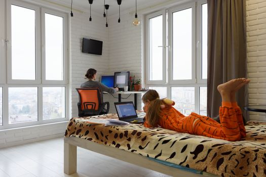 The family is at home, the child lies on the bed and studies in a laptop, the mother in the background works remotely in the computer