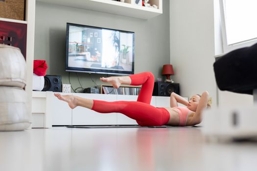 Attractive sporty woman working out at home, doing pilates exercise in front of television in small studio appartment. Social distancing. Stay healthy and stay at home during corona virus pandemic.