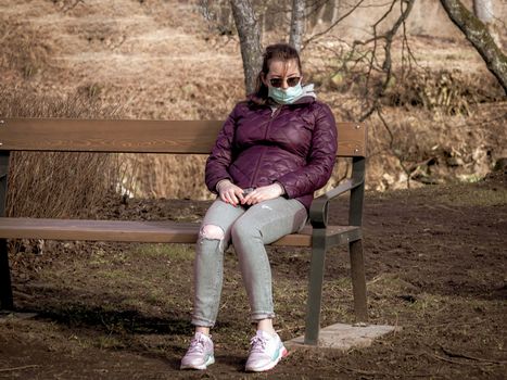 Lady and coronavirus protection. Sad Woman sitting alone on bench in park wearing mask to avoid infectious. Corona virus, or Covid 19, is spreading all over the world. Receives bad news on her phone.