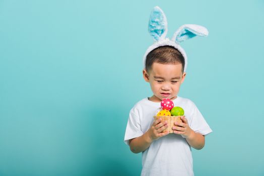 Asian cute little child boy smile beaming wearing bunny ears and a white T-shirt, standing to hold a basket with full Easter eggs on blue background with copy space