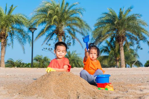 Asian Thai happy cute little cheerful Brother and sister two children funny digging play toy with sand at an outdoor tropical beach in summer day with copy space