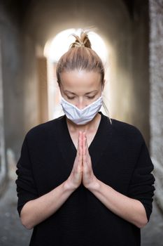 Coronavirus outbreak. Young caucasian woman wearing medical protection face mask praying over coronavirus global pandemic, for salvation of humanity, health, anxiety and depression reduction...