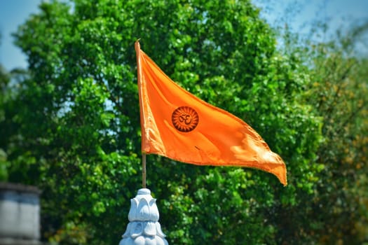 An orange flag containing a religious symbol in the center is waving at the peak of a temple. In the background green trees are present