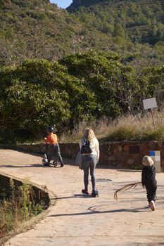 Mother and children walking on mountain path, 2 children, scooter, vegetation, bright sky