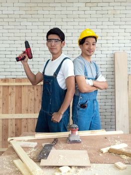 Two Asian carpentry poses confidently before beginning the job of receiving orders at the wood working place. Morning work atmosphere in the workshop room. A desk full of hand tools and wood piles.