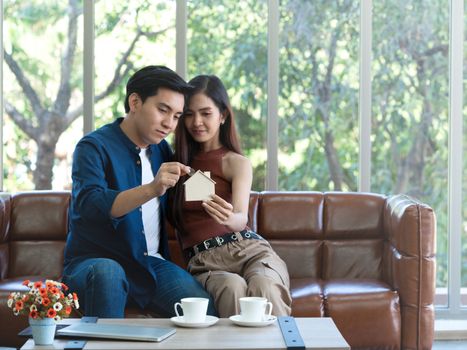 Young couples spend holidays on the leather sofa in the living room. The young man was wearing comfortable clothes, dropping the silver coins into a wooden house-shaped storing coin box. Focus on coin