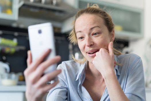 Young smiling cheerful woman indoors at home kitchen using social media apps on phone for video chatting and stying connected with her loved ones. Stay at home, social distancing lifestyle.