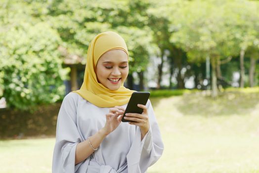 Portrait of cheerful Muslim girl using smartphone, smiling at outdoor.