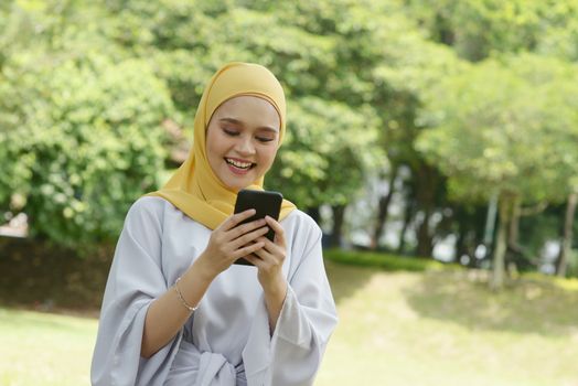 Portrait of cheerful Muslim girl using smartphone, smiling at outdoor.