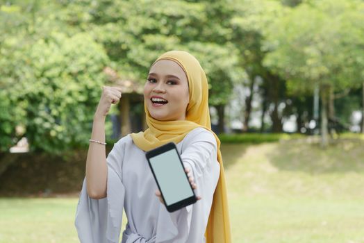 Portrait of cheerful Muslim girl using smartphone and thumb up, smiling at outdoor.