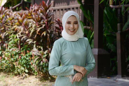 Portrait of Muslim girl in hijab, smiling at outdoor, traditional wooden house background. 