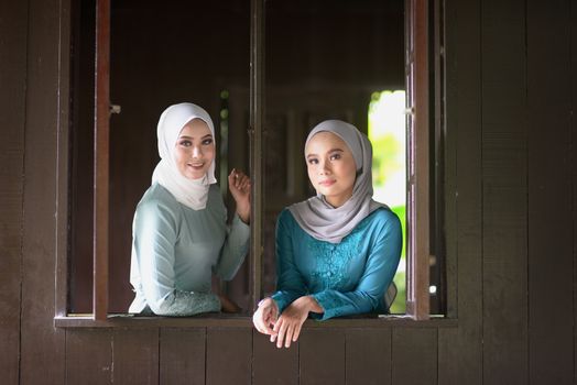 Portrait of Muslim girls in hijab smiling at traditional wooden house. 