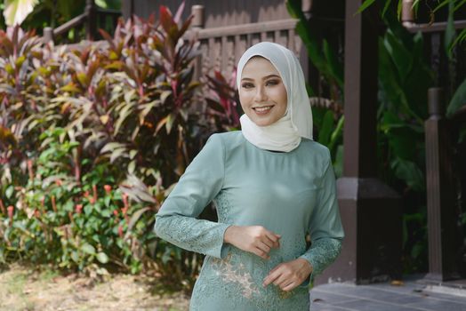 Portrait of Muslim girl in hijab, smiling at outdoor, traditional wooden house background. 