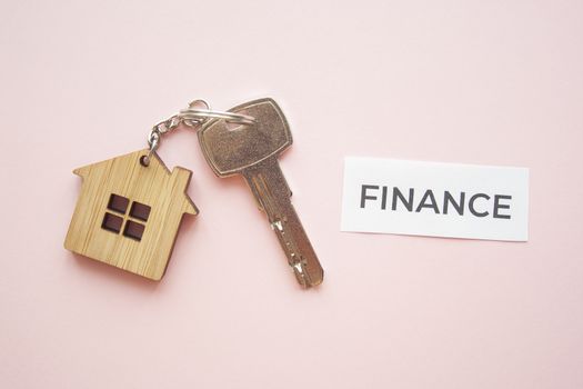 Wooden house toy and silver key on bright pink background with phrase quote Finance. Mortgage, house buy sell, investment, rent, realtor concept