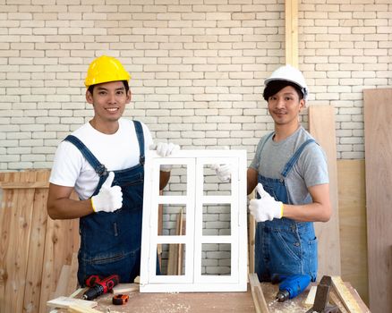 Two Asian carpenters proudly present the newly assembled window in his workshop. Handyman wearing a hard hat, Fabric gloves with a trusted drill In the morning work atmosphere