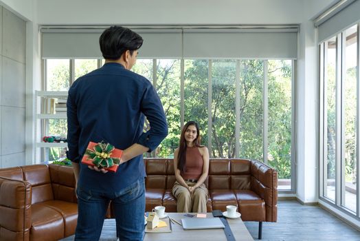 Young couples spend holidays in the living room. The young man wears comfortable clothes, hides a gift box behind, preparing to surprise girlfriend on special occasions.
