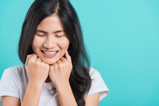 Portrait Asian beautiful happy young woman wear t-shirt glad keeps both hands under chin smiles pleasantly, on blue background, with copy space