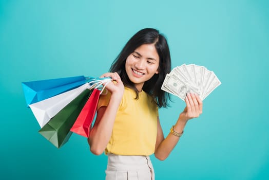 Portrait happy Asian beautiful young woman smile white teeth standing wear yellow t-shirt, She holding shopping bags and dollars money fan, studio shot on blue background with copy space for text