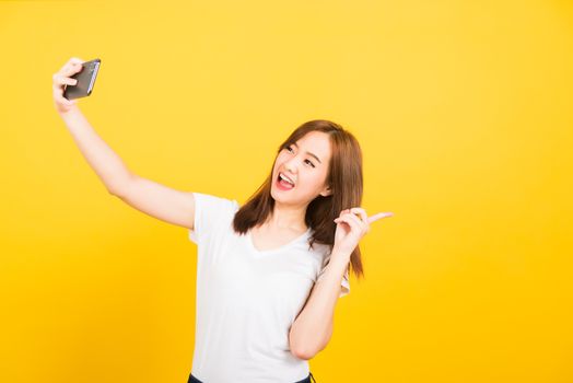 Asian happy portrait beautiful cute young woman teen smiling standing wear t-shirt mak selfie photo, video call on smartphone, point finger back isolated, studio shot yellow background with copy space