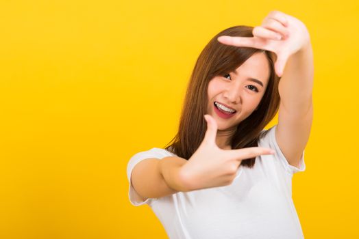 Asian happy portrait beautiful cute young woman standing wear t-shirt hands and finger making frame gesture camera for photography looking to camera, studio shot on yellow background with copy space