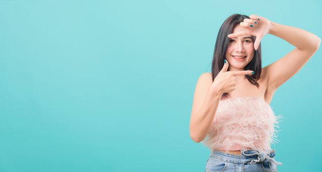 Asian happy portrait beautiful young woman standing smiling showing frame finger sign and looking to camera isolated on blue background with copy space for text