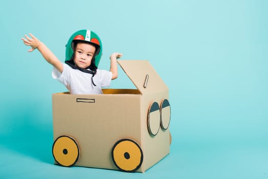 Happy Asian children boy with Helmet smile in driving play car creative by a cardboard box imagination, summer holiday travel concept, studio shot on blue background with copy space for text