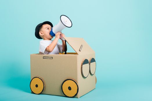 Happy Asian children boy smile in driving play car creative by a cardboard box imagination with megaphone, summer holiday travel concept, studio shot on blue background with copy space for text