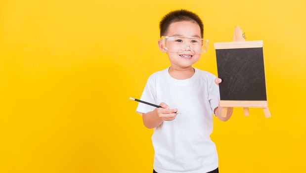 Asian Thai happy portrait cute little cheerful child boy smile he showing blackboard and looking to camera, studio shot isolated on yellow background with copy space