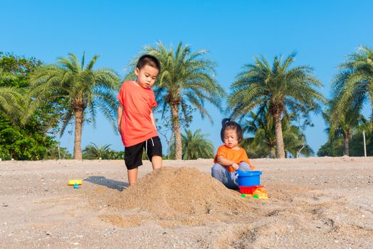 Asian Thai happy cute little cheerful Brother and sister two children funny digging play toy with sand at an outdoor tropical beach in summer day with copy space