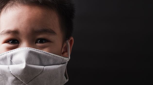 Closeup Asian little child wearing protective face mask with fear in the eye, prevent germs or disease hygiene prevention COVID-19 virus or coronavirus protection concept, dark on black background