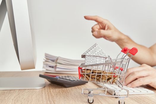 House on gold coins in shopping cart have blur man pointing finger for checking sales of shopping online concept and pile report of sale on wooden computer table with white background and copy space.