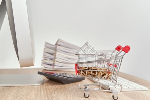 House on gold coins in shopping cart have blur calculator with pencil and pile report of sale on wooden computer table with white background and copy space. Business concepts shopping online.