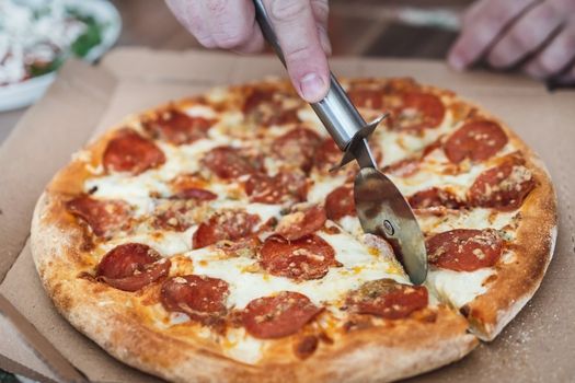 cropped male hand cuts pizza margarita with pepperoni with a special knife, close-up.