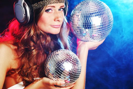 Disco girl enjoys music in head phones holding mirror disco ball over black and colorful smoke background