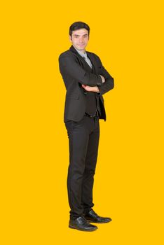 A young businessman in a gray shirt and a black suit fold his arms over the chest with confidence. The concept of investing in a successful business. Portrait on a yellow background with studio lights