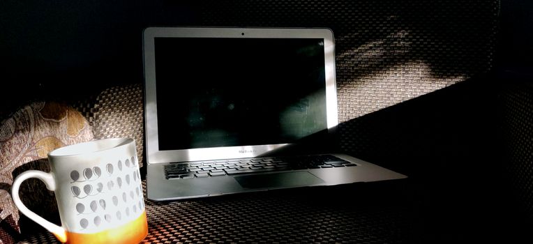 Screen of a laptop kept on the couch with a steaming cup of coffee basking in the rays of sun in the morning