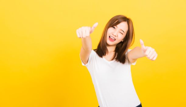 Asian happy portrait beautiful cute young woman teen standing wear t-shirt showing gesturing finger thumb up looking to camera isolated, studio shot on yellow background with copy space