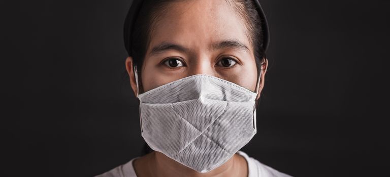 Closeup Asian young woman wearing protective face mask with fear in the eye, prevent germs or disease hygiene prevention COVID-19 virus or coronavirus protection concept, dark on black background