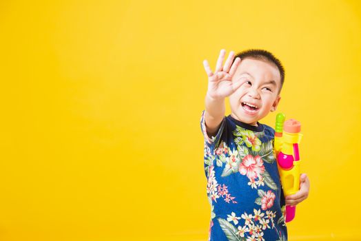 Portrait happy Asian cute little children boy smile standing so happy wearing flower shirt in Songkran festival day holding water gun, studio shot on yellow background with copy space
