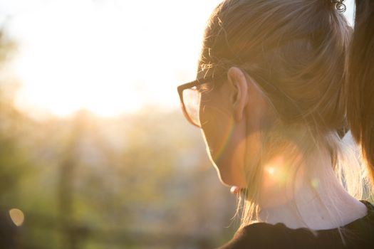 Backlit rear view of young woman talking on cell phone outdoors in park at sunset. Girl holding mobile phone, using digital device, looking at setting sun. Copyspace.