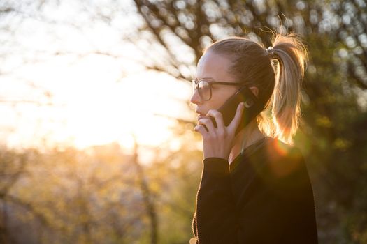Backlit rear view of young woman talking on cell phone outdoors in park at sunset. Girl holding mobile phone, using digital device, looking at setting sun. Copyspace.