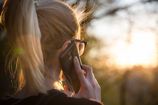 Backlit rear view of young woman talking on cell phone outdoors in park at sunset. Girl holding mobile phone, using digital device, looking at setting sun. Copyspace.