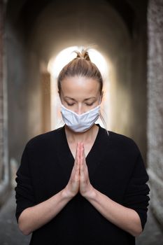 Coronavirus outbreak. Young caucasian woman wearing medical protection face mask praying over coronavirus global pandemic, for salvation of humanity, health, anxiety and depression reduction...