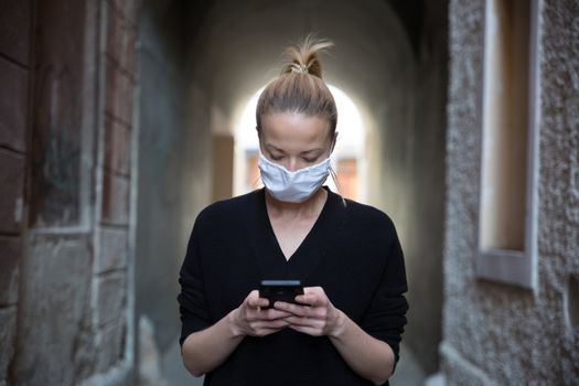 COVID-19 pandemic coronavirus. Casual caucasian woman at medieval city street using mobile phone, wearing protective face mask against spreading of coronavirus and disease transmission.
