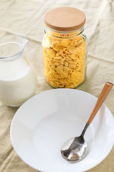 Cornflakes in a jar, next to a transparent jug of milk, an empty plate with a spoon on a plain rough tablecloth. View from above