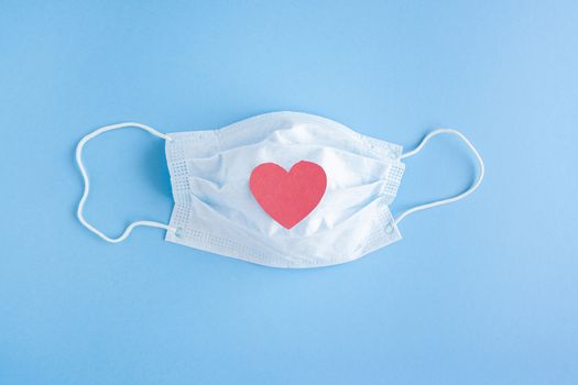 Disposable medical mask on blue background with red heart on it against bacteria and viruses. Stop the spread of coronavirus, self-defense. Copy space