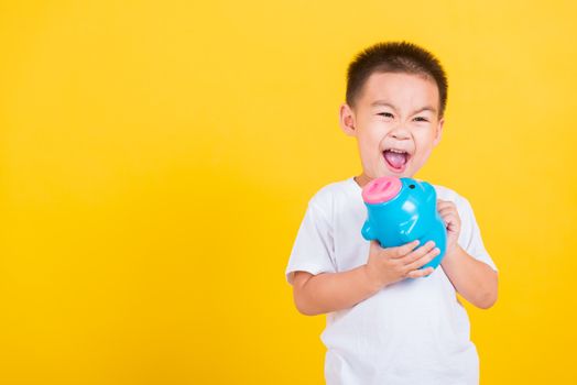 Asian Thai happy portrait cute little cheerful child boy smile putting coin money to the piggy bank and looking camera, studio shot isolated on yellow background with copy space