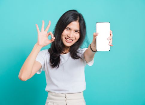 Portrait Asian beautiful happy young woman standing smile, holding mobile phone and showing ok gesture, shoot photo in studio focus phone on blue background, with copy space