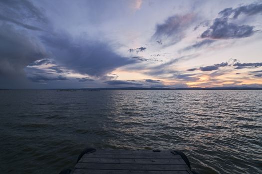 Landscape of a lake with storm clouds, moving waters reeds in the water, bluish tones, pier